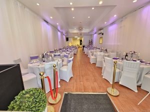 his photo showcases a long, elegantly decorated event hall with white drapes, warm wood flooring, and purple lighting accents. Round tables are perfectly set with floral centerpieces, candles, and sparkling tableware, creating a memorable setting for guests.