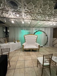 Elegant gold and white throne chair with floral arch backdrop, perfect for weddings, bridal showers, and milestone celebrations in Queens.