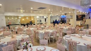 Wide view of a fully decorated banquet hall featuring blush pink accents, floral centerpieces, and crystal chandeliers, ideal for weddings, birthdays, and special occasions at Queens Party Hall.