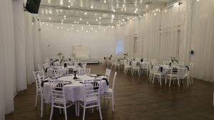A bright, airy event hall featuring white Chiavari chairs, round tables with black and white table settings, sheer draped walls, and warm string lighting. Ideal for weddings, receptions, and corporate events in Queens.